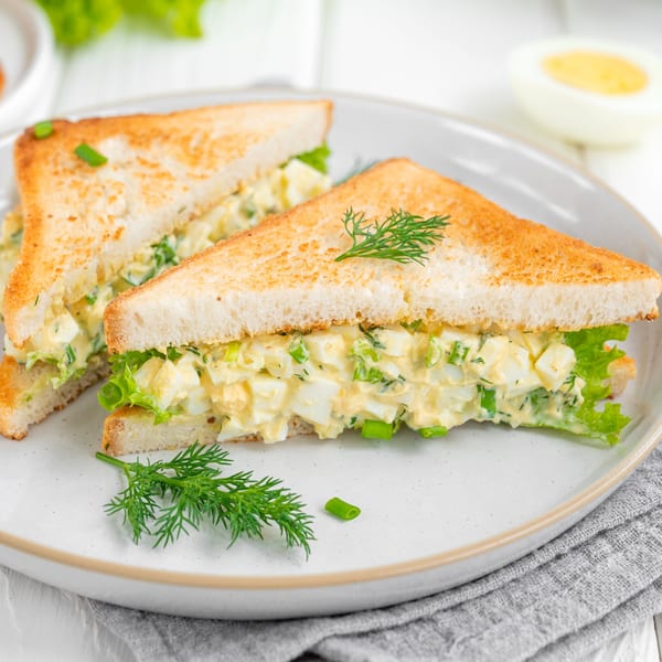 Two egg salad sandwiches on toasted bread with lettuce, dill, and green onions on a white plate.
