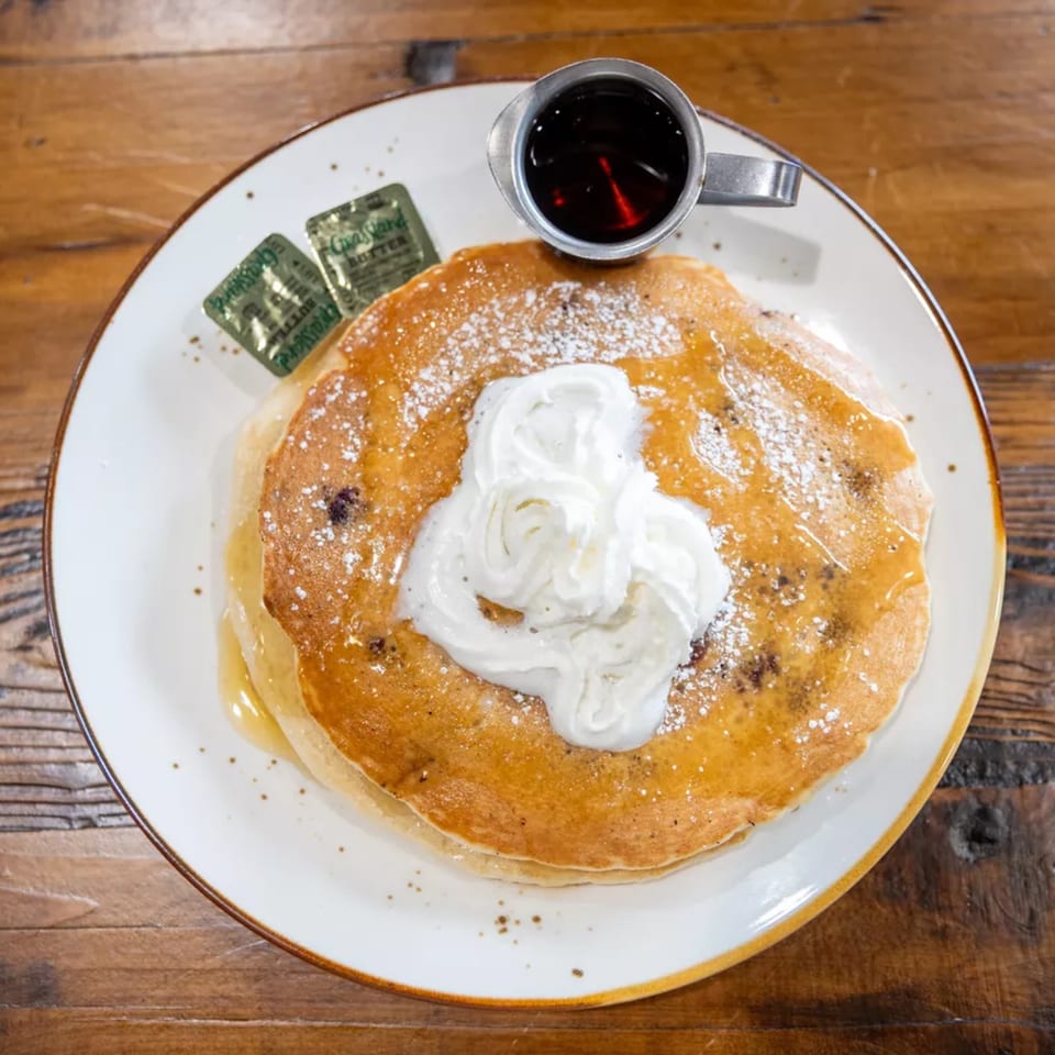 Golden pancakes topped with whipped cream and powdered sugar, served with syrup in a metal cup on a white plate.