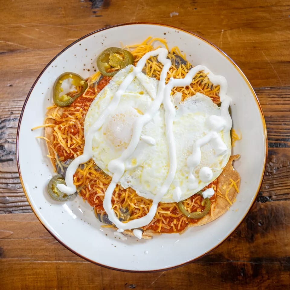 Plate of nachos topped with fried eggs, shredded cheese, jalapeños, red sauce, and sour cream on a wooden table.