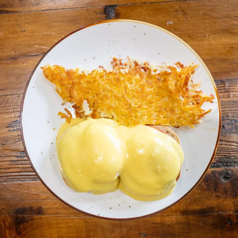 Plate of eggs Benedict with hollandaise sauce and crispy hash browns on a rustic wooden table.