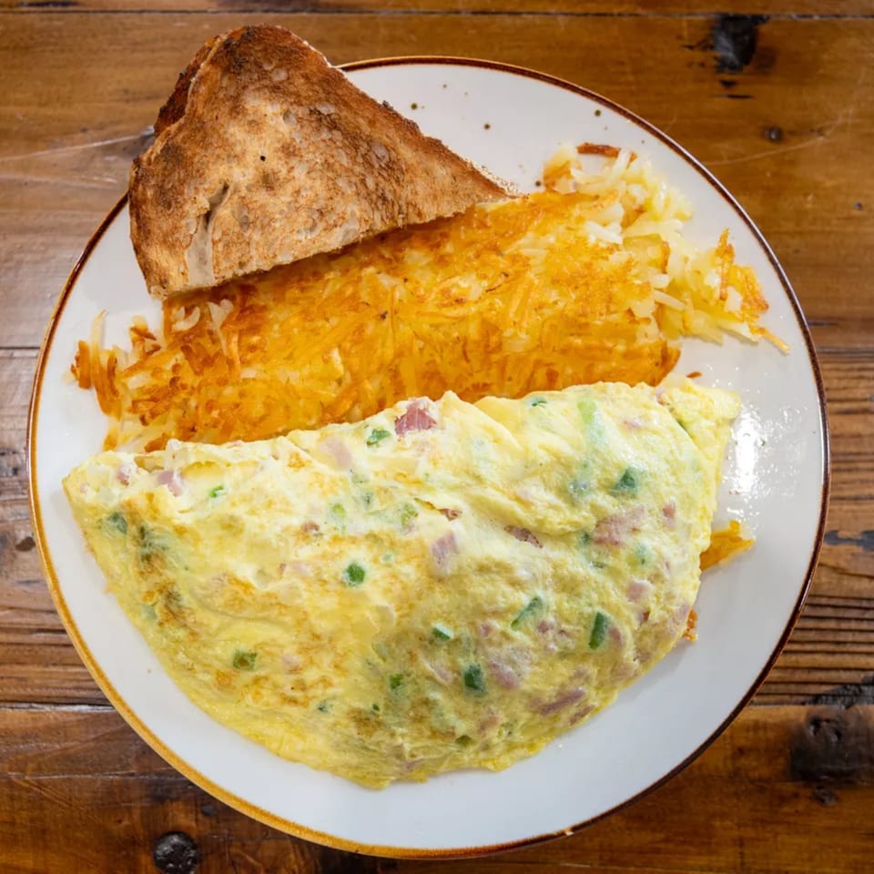 Cheesy folded omelet with diced ham and green peppers, shredded hash browns, and toasted multigrain bread on a plate.
