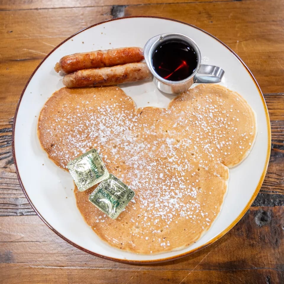 Plate of powdered-sugar pancakes with butter packets, two sausages, and syrup on a wooden table.